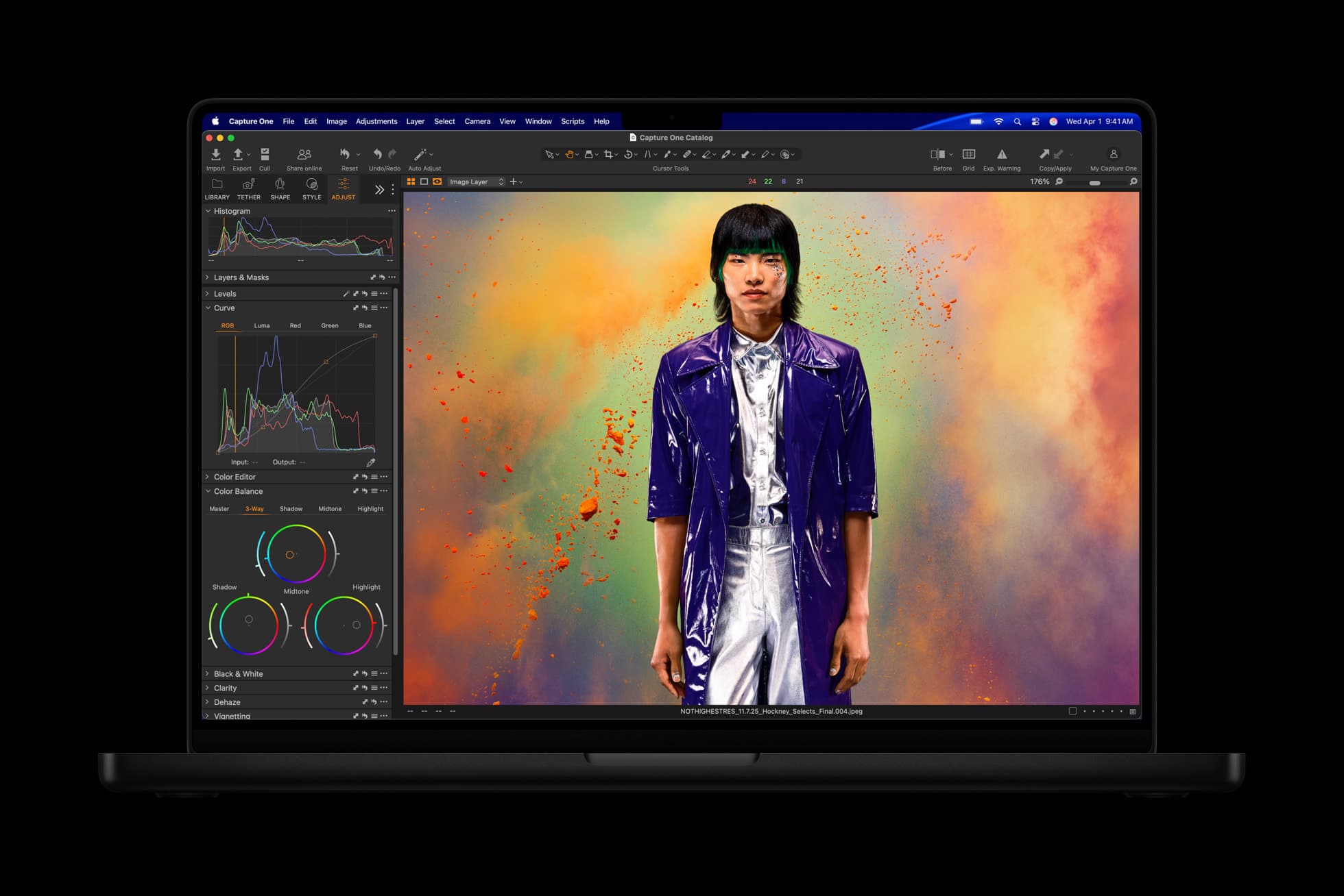 A space black MacBook Pro seen head-on against a black background, its screen showing a photo-editing window with histogram and color-wheel panels on the left and, on the right, a person in a glossy purple coat and silver shirt standing in a cloud of orange and green powder.