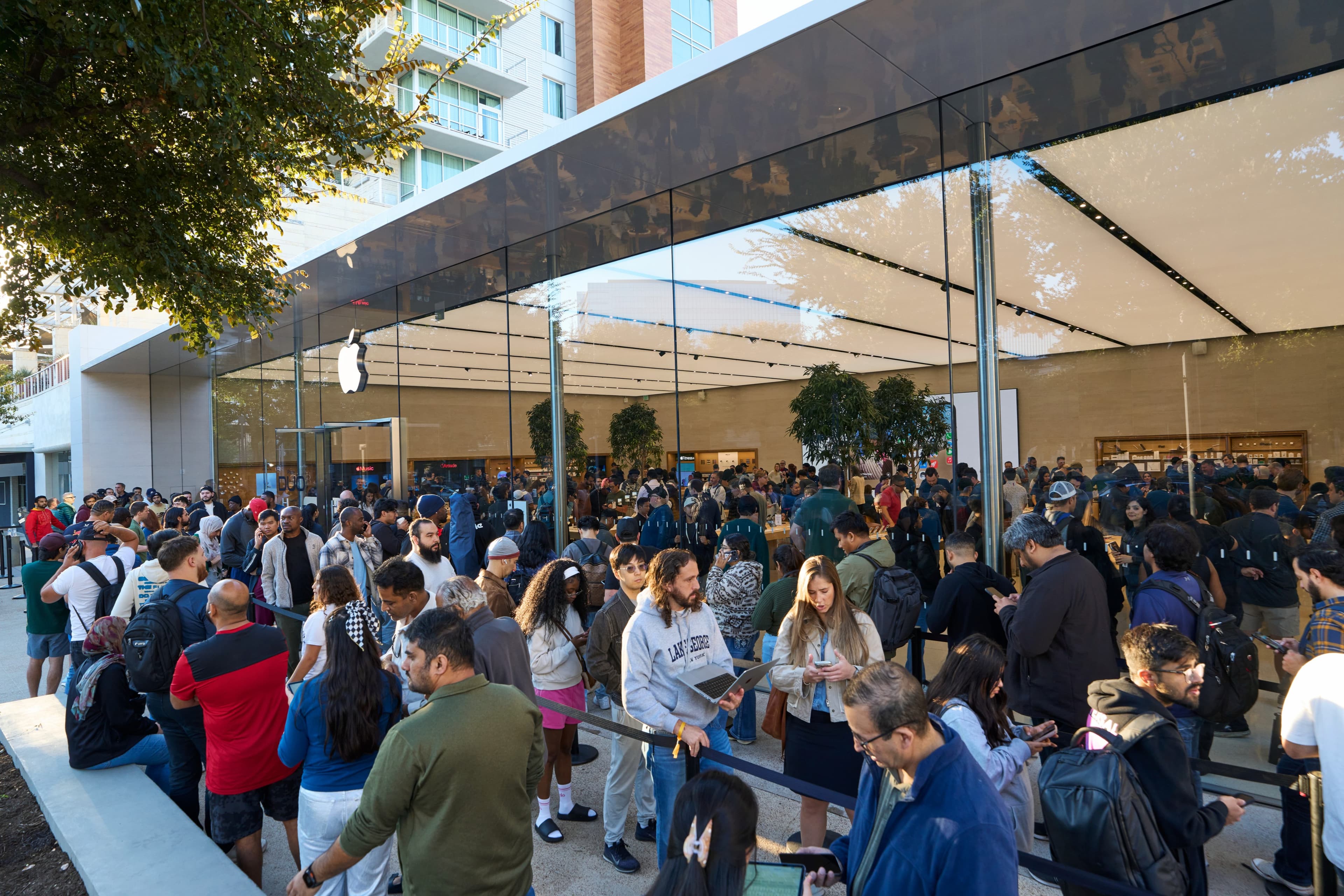 A crowd waiting behind a rope line outside the glass-walled Apple Domain NORTHSIDE store in Austin, Texas, in morning sun, most of them looking at their phones and one holding an open MacBook, with the Apple logo on the glass, trees overhead and a tall apartment building behind the store.