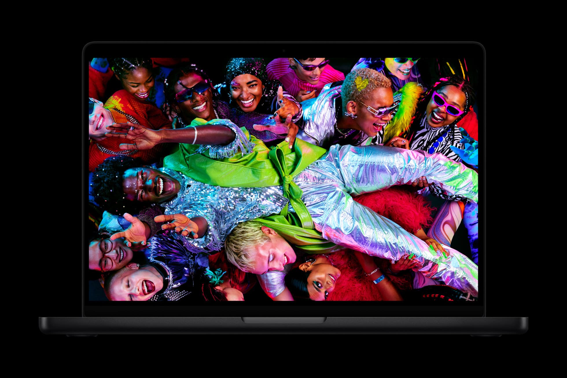 A space black MacBook Pro seen head-on against a black background, its lid open and its screen filled with a photograph of a laughing crowd in sequins, metallic silver and neon green, carrying one person stretched across their raised hands.